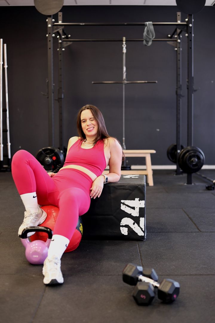 Julieta in pink sportswear in the studio, leaning on a training box, winking and sticking her tongue out