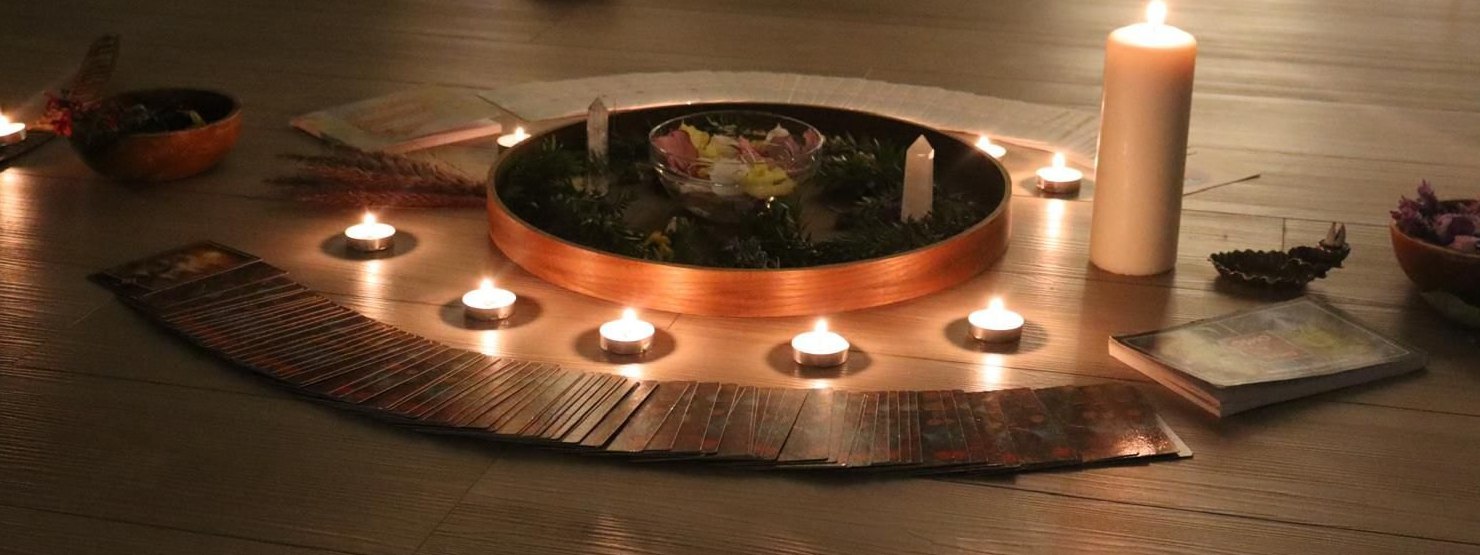 מעגל של נרות דולקים סביב מגש נחושת עם עלים, קריסטלים ועלי ורדים, וקלפים פרושים בחצי עיגול על הרצפה
