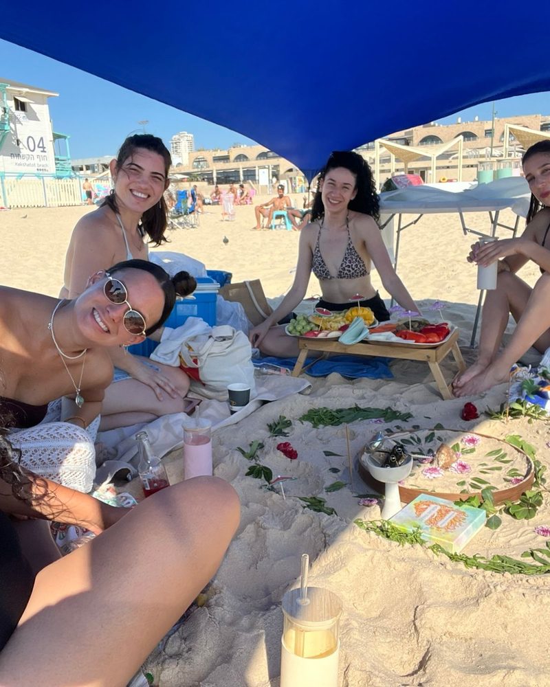 חברות יושבות בחוף סביב מעגל של עלים ופרחים בחול