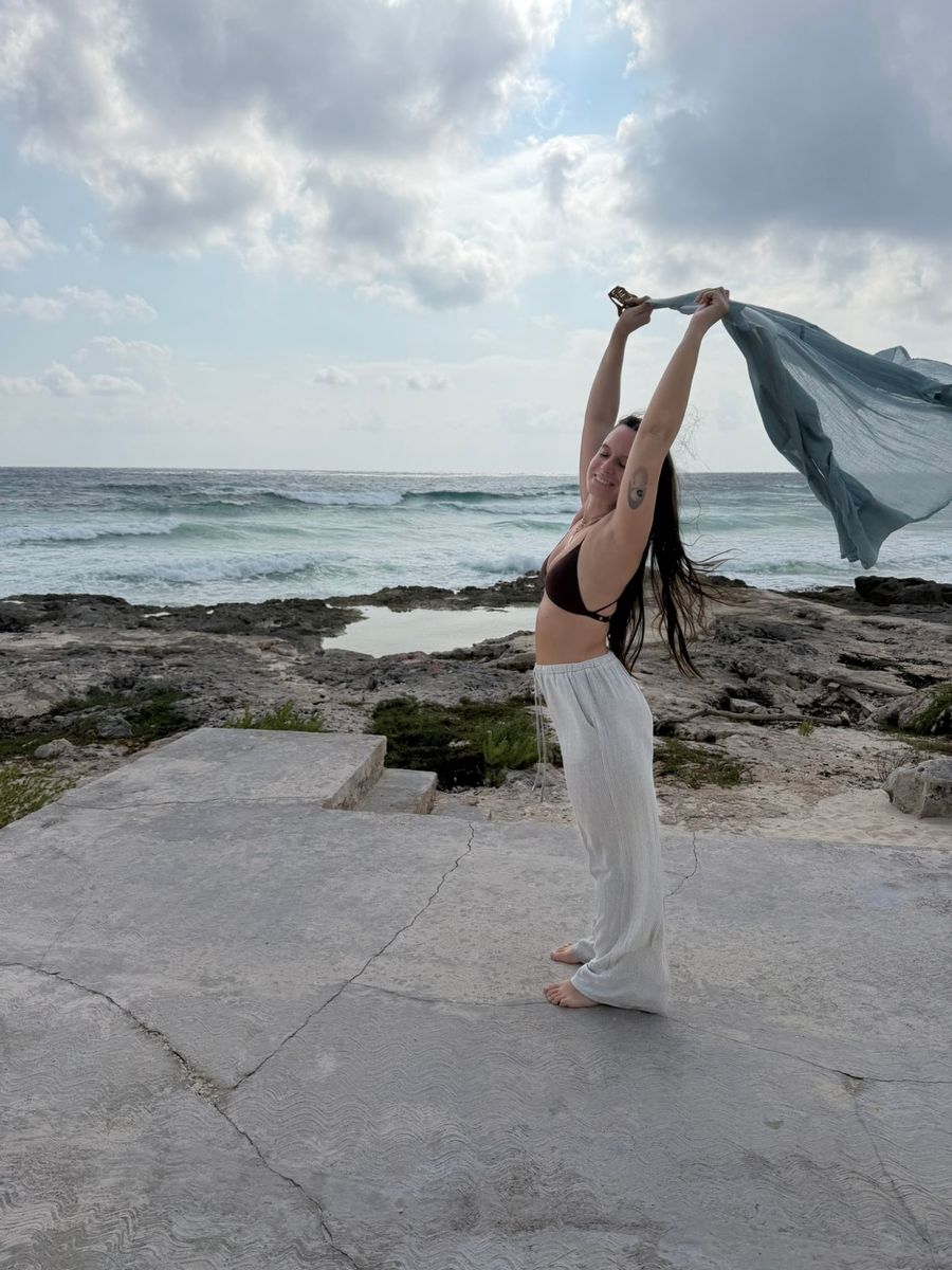 Julieta on a rock by the sea, eyes closed and smiling, holding a light-blue cloth over her head as it flutters in the wind
